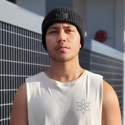 A man wearing a white sleeveless tank top with a small black geometric star logo on the chest, paired with a black knit beanie and blue jeans, standing outdoors in front of a modern building with a metal grid fence.