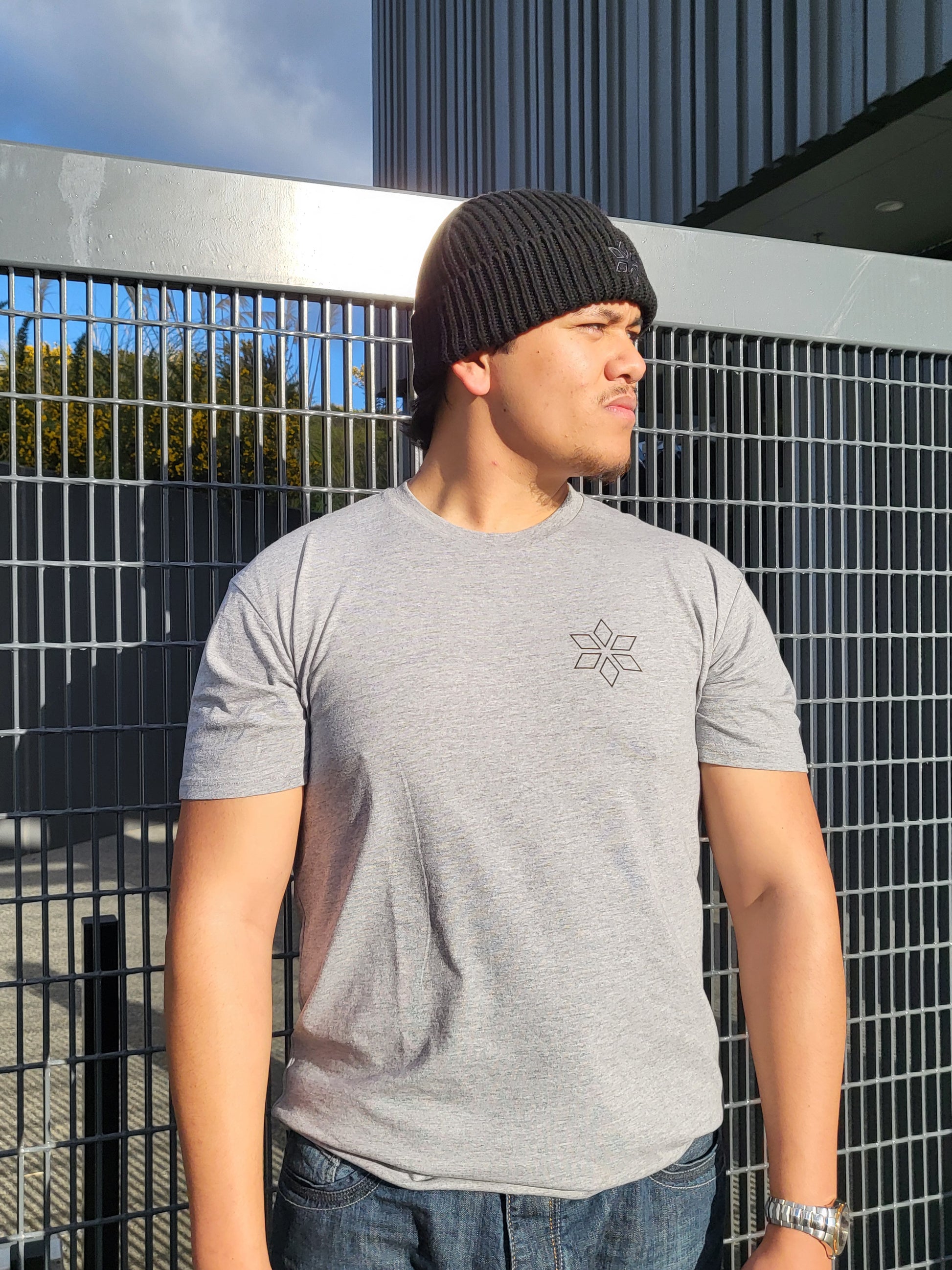 A man wearing a light gray t-shirt with a small geometric star logo on the chest, paired with a black beanie and blue jeans, leaning against a metal grid fence.