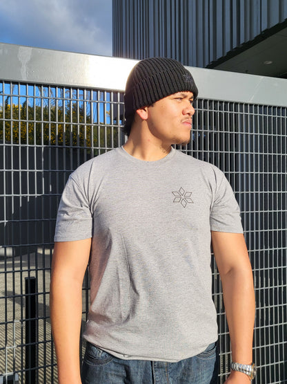 A man wearing a light gray t-shirt with a small geometric star logo on the chest, paired with a black beanie and blue jeans, leaning against a metal grid fence.