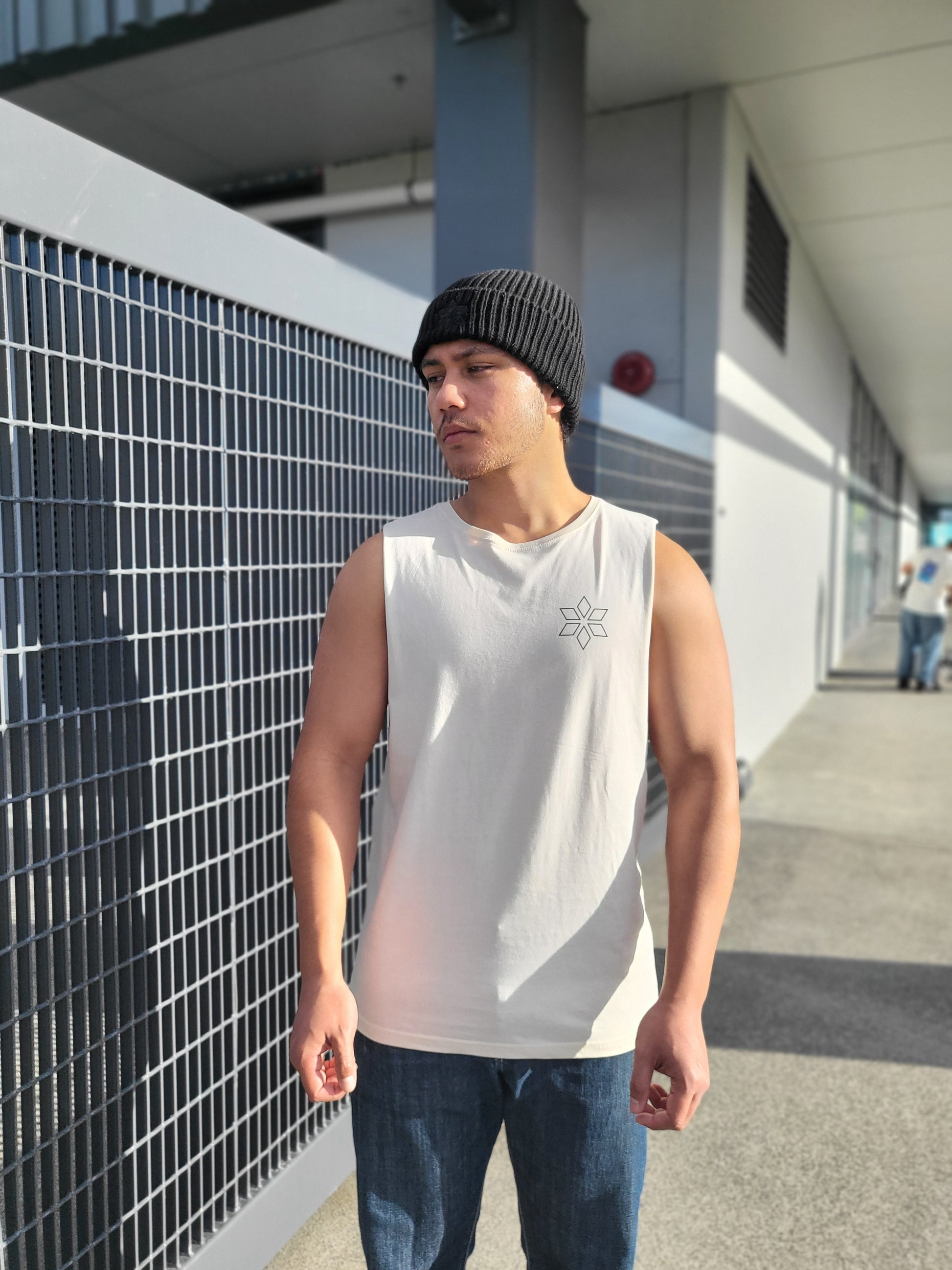 A man wearing a cream sleeveless tank top with a small black geometric logo on the left chest, blue jeans, and a black knit beanie, standing outdoors next to a metal grid fence.