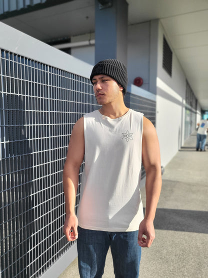 A man wearing a cream sleeveless tank top with a small black geometric logo on the left chest, blue jeans, and a black knit beanie, standing outdoors next to a metal grid fence.
