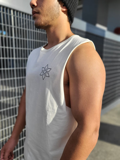 A man wearing a cream sleeveless tank top with a black geometric line-art logo on the left chest, photographed outdoors against a metal grid fence.