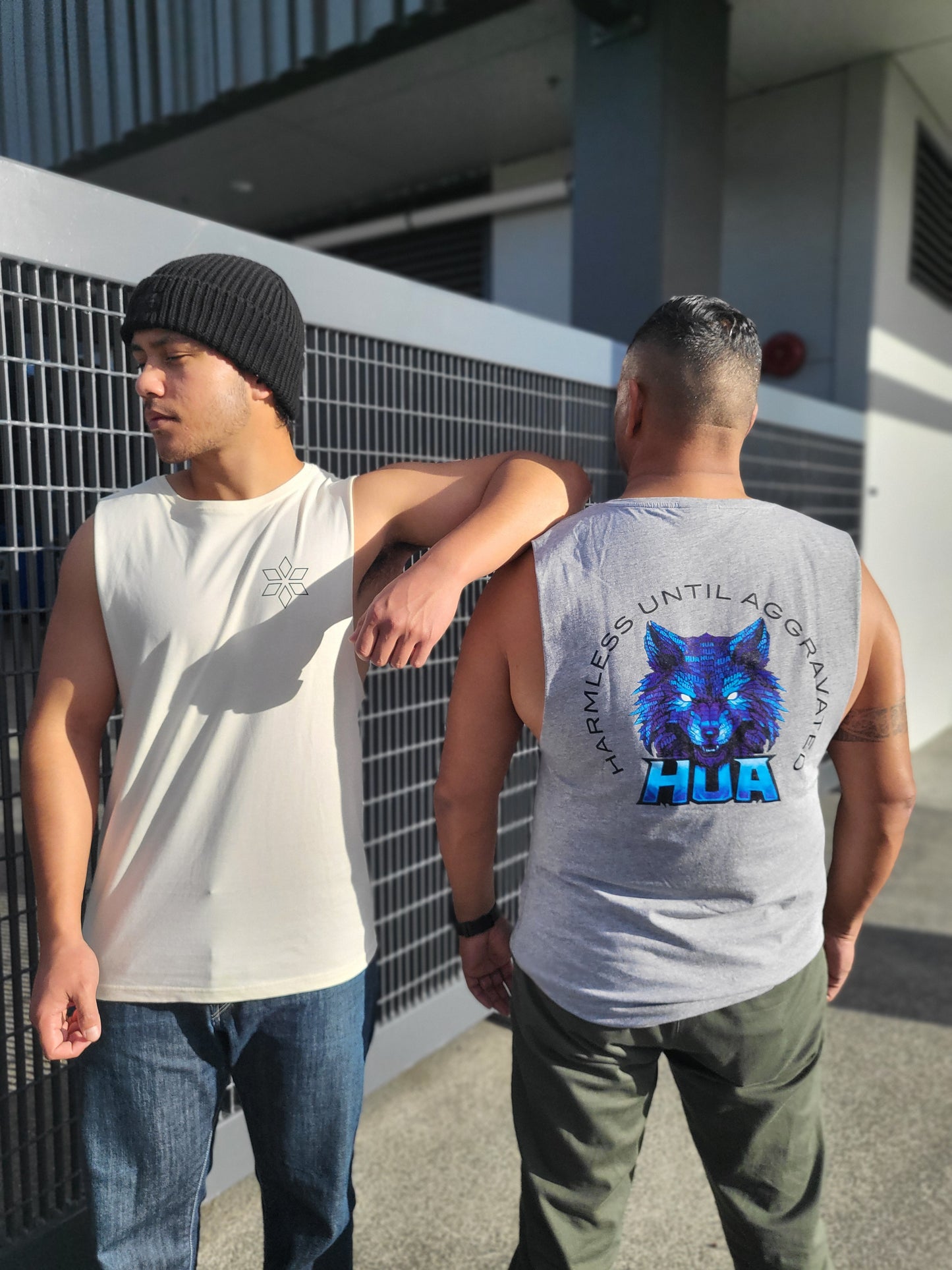 Two men standing outdoors next to a metal fence, showcasing sleeveless tank tops. The man on the left wears a cream tank top with a small black star logo on the chest, paired with a black beanie.